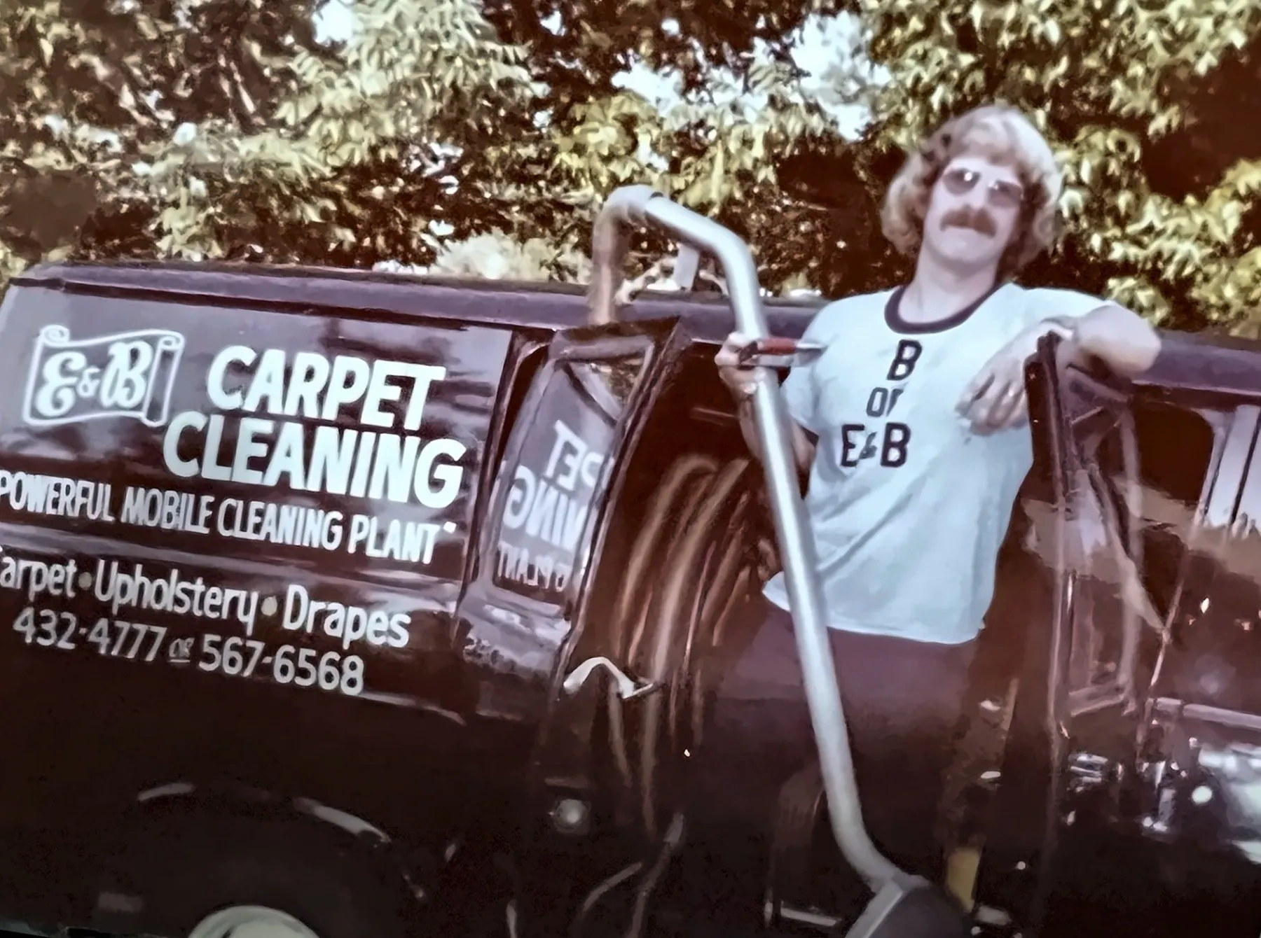Brian, the B of E&B, in his B of E&B T-shirt leaning on the open door of the black E&B Carpet Cleaning van with a cleaning wand, 1970s