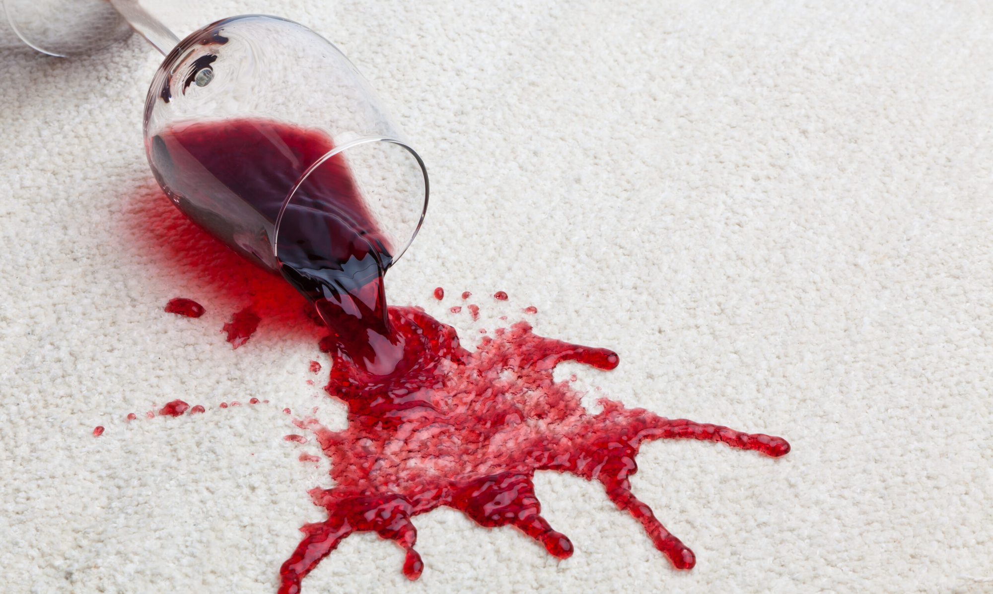 A tipped glass spilling red wine onto a cream carpet